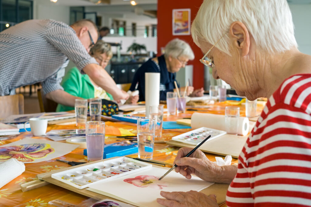 Woman in striped red and white shirt working on watercolor painting at table with other students in spacious studio.